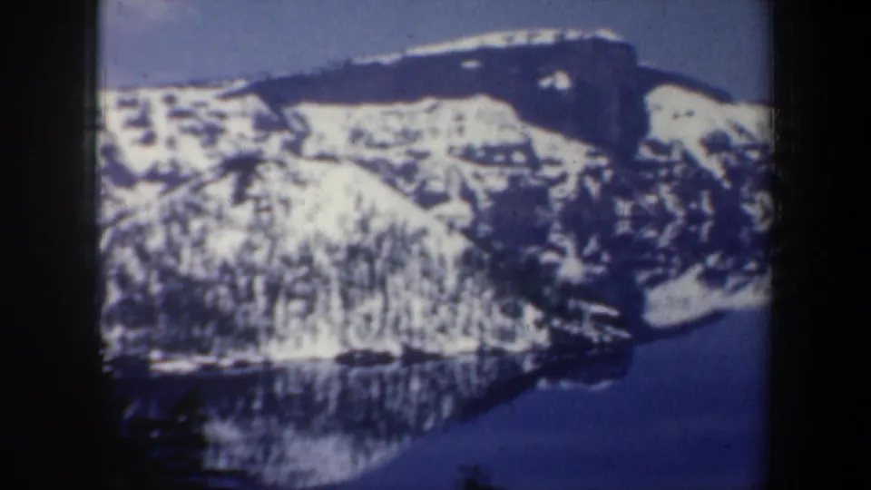 1957 Oregon 8mm frame showing snowy mountain slopes reflected in a dark lake, with bare and evergreen trees in front.