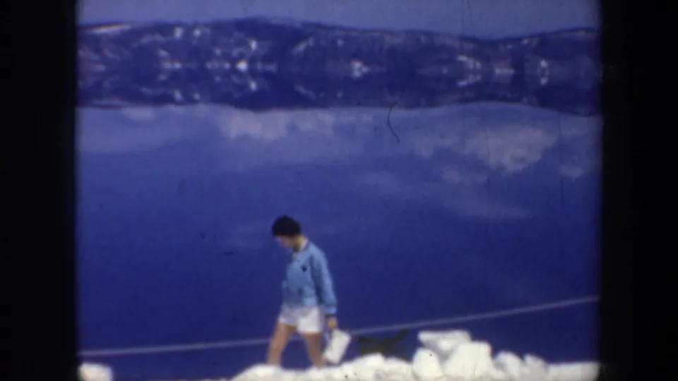 1957 Oregon 8mm frame showing a woman in a blue sweater walking over icy snow while carrying a small white bag.