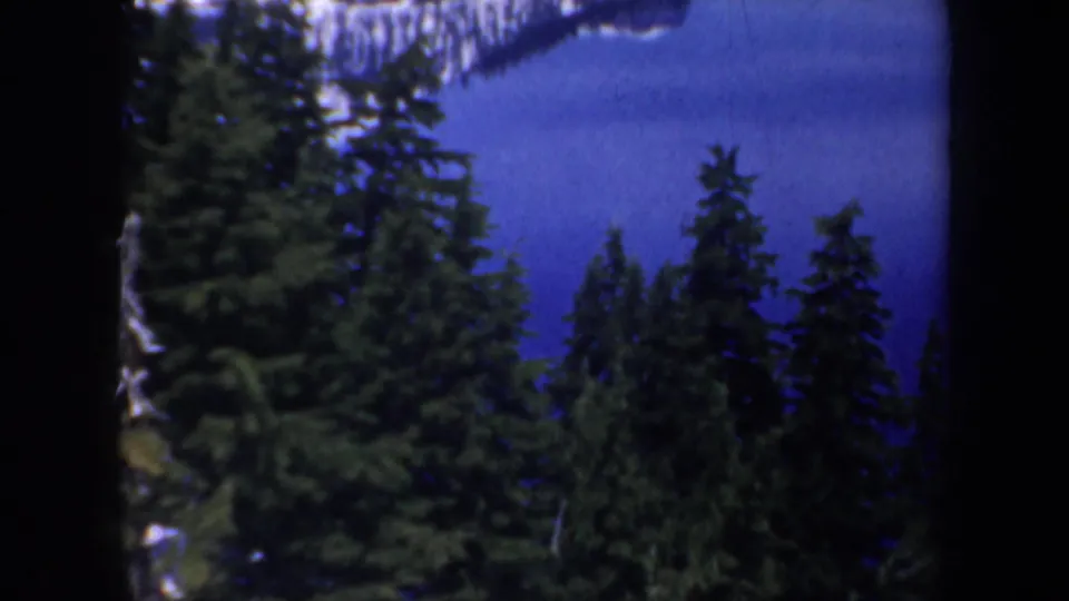 1957 Oregon home-movie frame with conifers, blue lake water, and a pale mountain reflection beyond the trees.