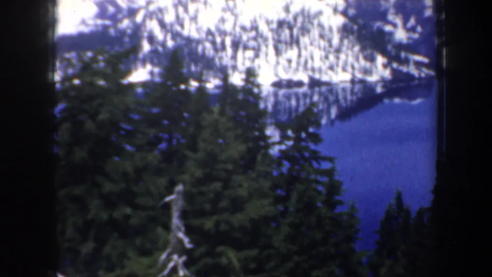 1957 Oregon 8mm frame showing foreground conifers and a blue lake reflecting snow-covered mountains.