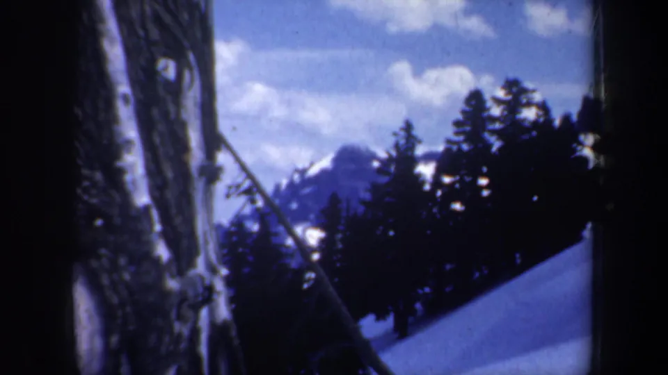 1957 Oregon 8mm frame looking past tree trunks and evergreens toward a snowy slope and distant mountain peak.