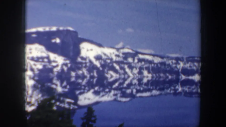 1957 Oregon home-movie view of snow-covered mountains above a lake, framed by dark foreground trees and soft 8mm color.