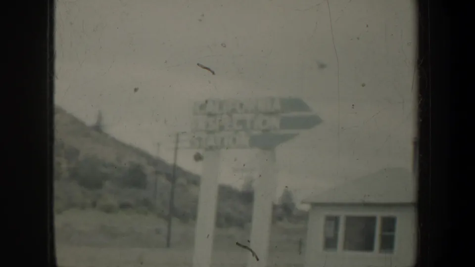 Soft, faded 8mm frame of a roadside inspection-station sign, nearby building, utility poles, and low hills during travel.