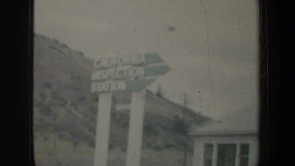 California Inspection Station sign fills the roadside view, with a small building, hillside, and utility poles behind it in 1950s 8mm footage.