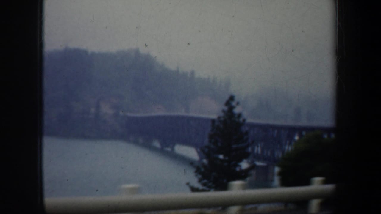 1957 Oregon 8mm home-movie frame showing a distant steel truss bridge over water with wooded hills beyond, partly obscured by dark window framing and a roadside rail.
