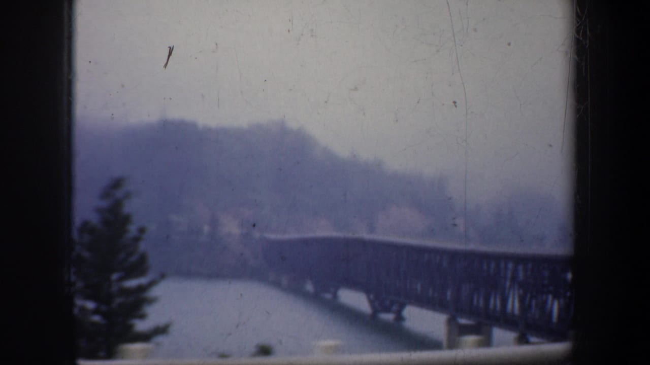 1957 Oregon 8mm frame showing a steel truss bridge spanning water below wooded hills, with the view partially divided by dark vehicle-window framing.