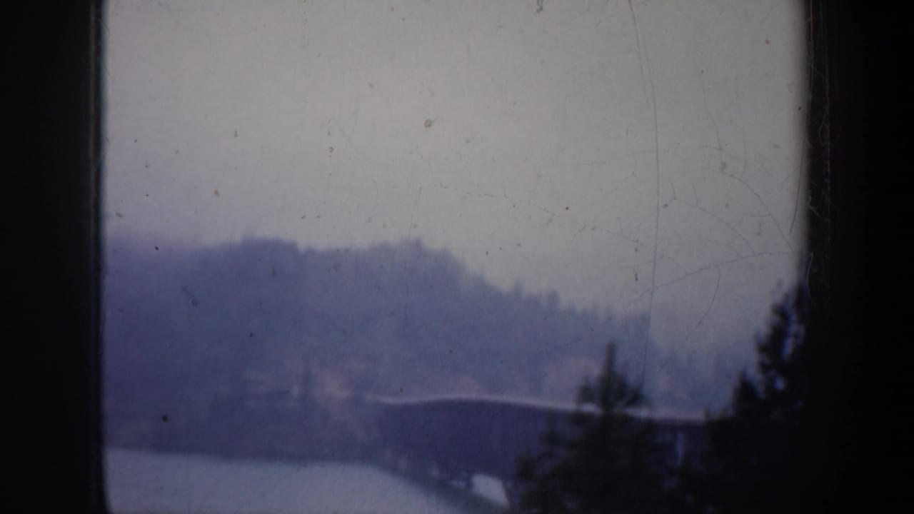 Soft 1957 Oregon home-movie view of a bridge crossing water beneath hazy hills, framed by dark foreground edges and roadside elements from a moving car.