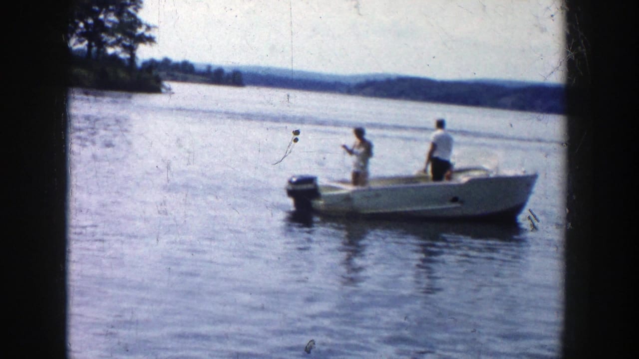 Two men stand in a small motorboat on a lake, with shoreline and trees in the distance.
