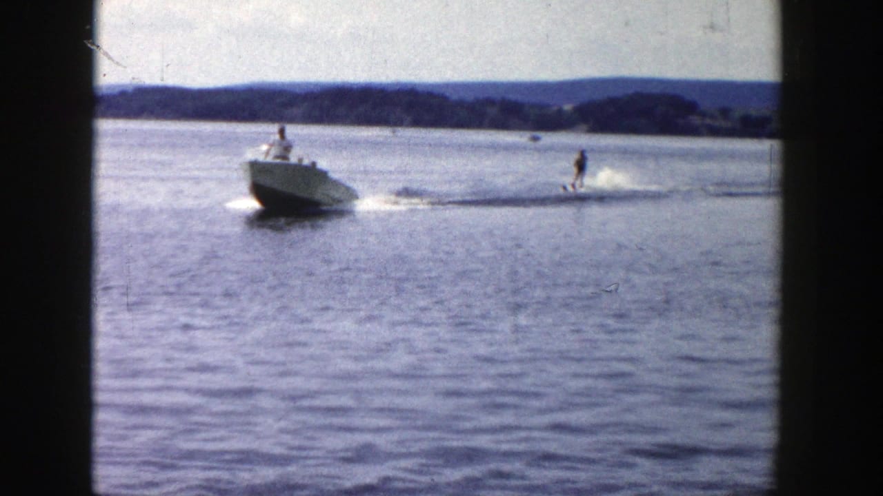 1958 Wisconsin water-skiing scene with a white ski boat pulling a skier across open lake water, with hills in the distance.