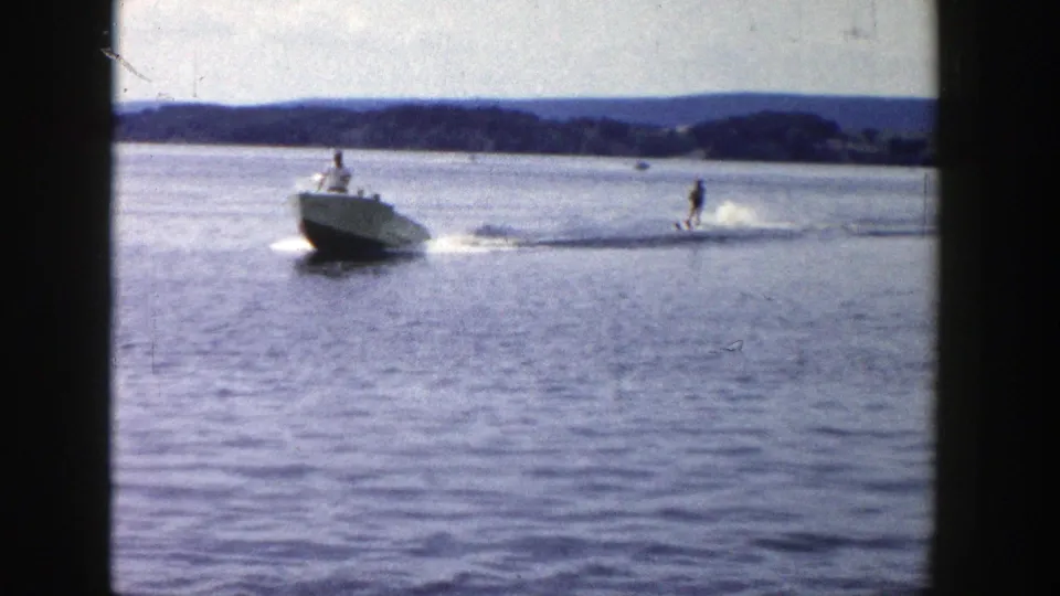 1958 Wisconsin water-skiing scene with a white ski boat pulling a skier across open lake water, with hills in the distance.