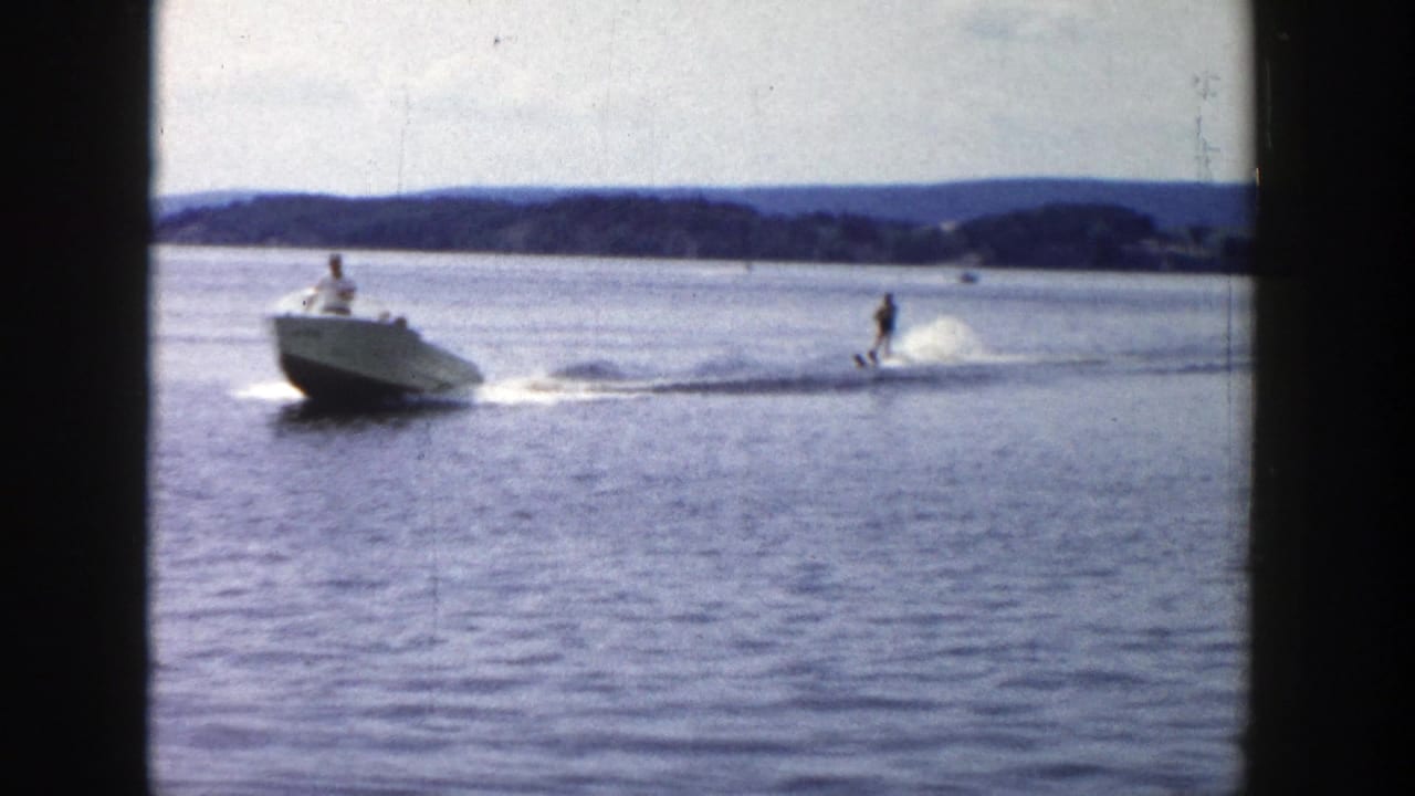 1958 Wisconsin 8mm frame showing a small motorboat towing a water skier on a calm lake with low wooded hills in the distance.