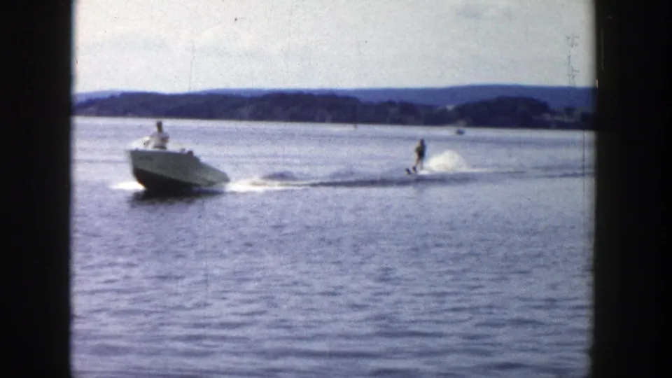 1958 Wisconsin 8mm frame showing a small motorboat towing a water skier on a calm lake with low wooded hills in the distance.