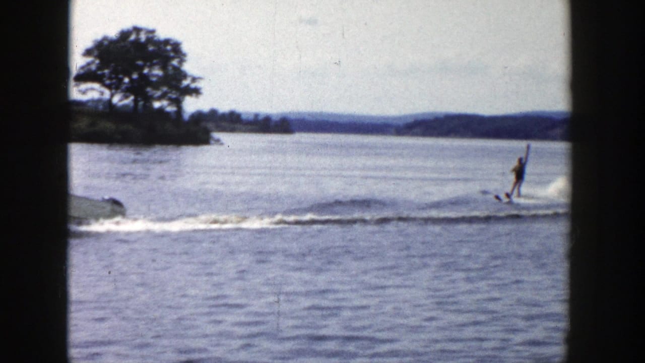 1958 Wisconsin 8mm frame with a water skier upright behind a motorboat, with trees and shoreline visible across the lake.