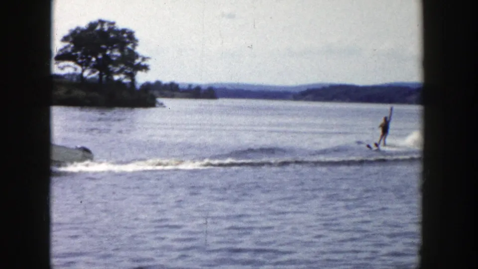 1958 Wisconsin 8mm frame with a water skier upright behind a motorboat, with trees and shoreline visible across the lake.