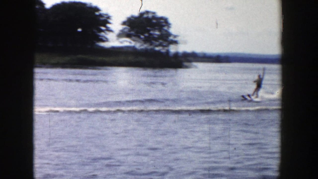 1958 Wisconsin home-movie frame showing a distant water skier moving across calm lake water near a wooded shoreline.
