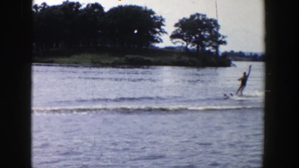 1958 Wisconsin home-movie frame of a distant man water skiing across a calm lake behind dark window framing.