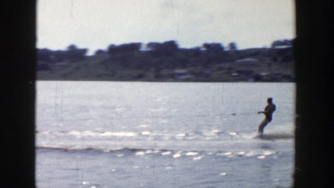 The skier leans back while crossing the lake, with low shoreline and distant structures softened by 8mm film grain.