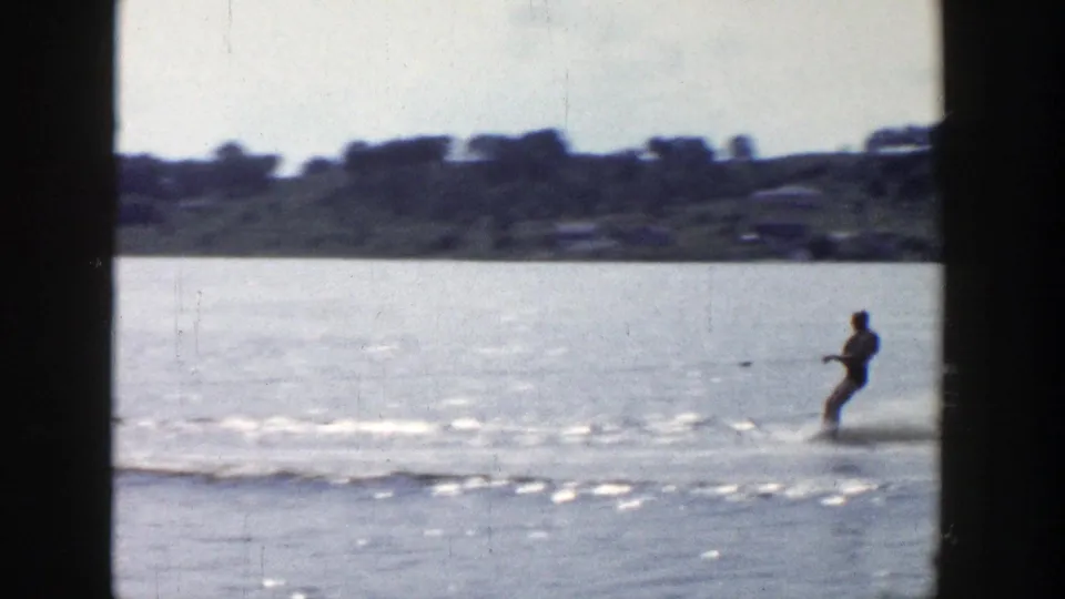 The skier leans back while crossing the lake, with low shoreline and distant structures softened by 8mm film grain.