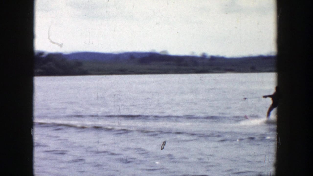 Near the end of the shot, the skier moves out of frame across open lake water with low wooded hills in the distance.