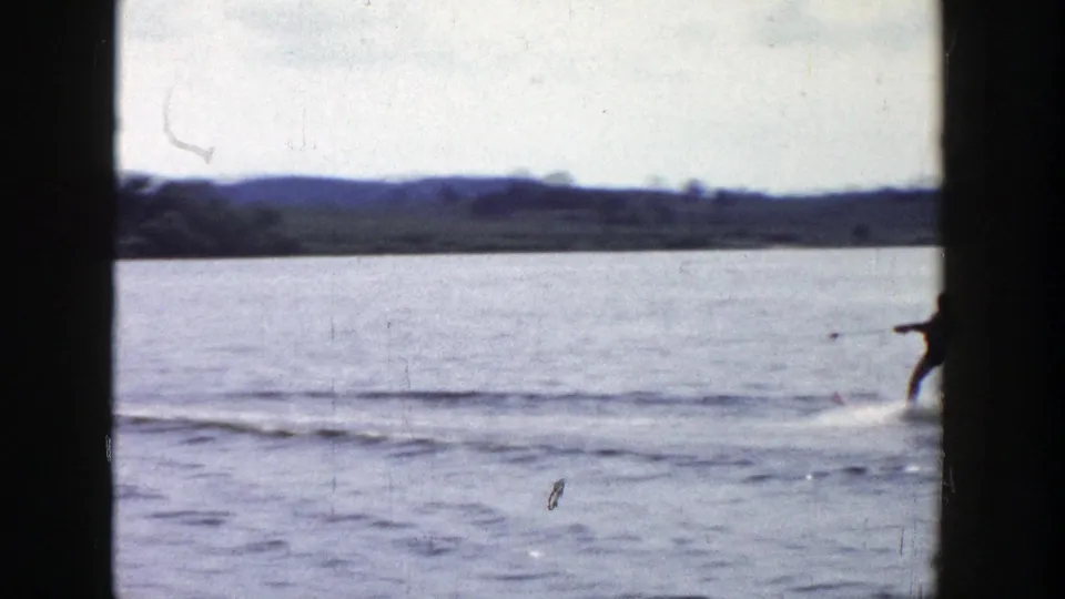 Near the end of the shot, the skier moves out of frame across open lake water with low wooded hills in the distance.