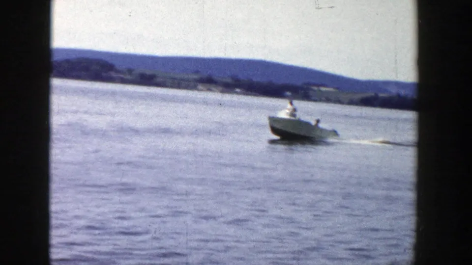 1958 Wisconsin 8mm frame of a small motorboat pulling a water skier across a lake with wooded shoreline and low hills beyond.