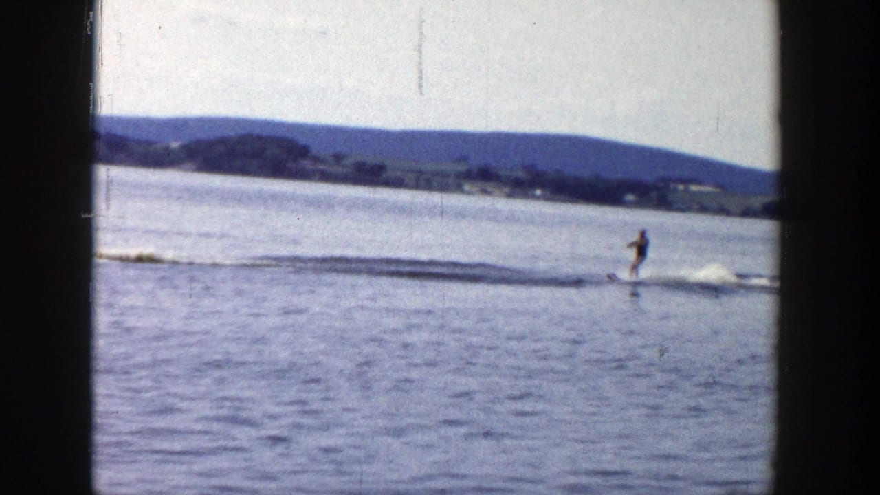 A water skier in dark swimwear rides across the lake surface behind a motorboat wake in 1958 Wisconsin home-movie footage.