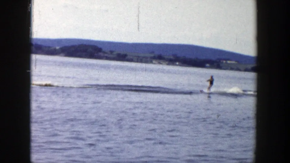 A water skier in dark swimwear rides across the lake surface behind a motorboat wake in 1958 Wisconsin home-movie footage.