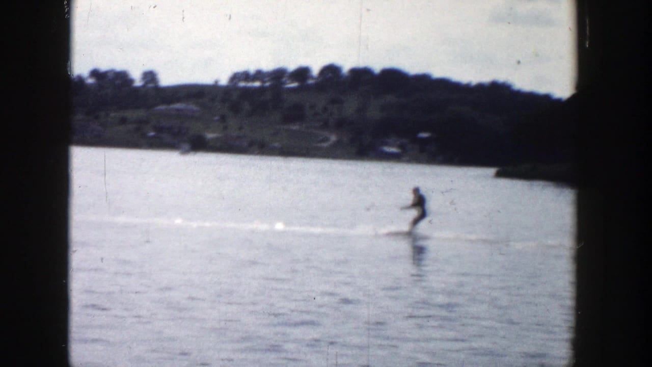 In 1958 Wisconsin home-movie footage, a distant water skier moves across a calm lake with shoreline trees behind.