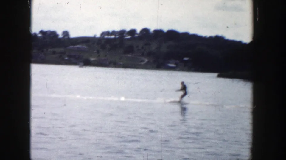 In 1958 Wisconsin home-movie footage, a distant water skier moves across a calm lake with shoreline trees behind.