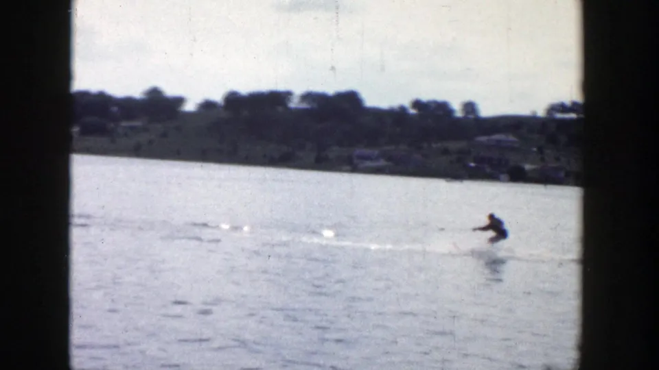 A distant skier in 1958 Wisconsin footage leans across the water while a white wake cuts over the lake surface.