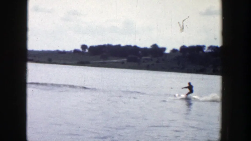 The 8mm frame shows a skier skimming over a Wisconsin lake, with a low wooded shoreline and soft sky in the background.