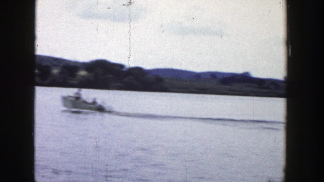 1958 Wisconsin 8mm frame showing a small motorboat crossing calm lake water with a wooded, hilly shoreline in the distance.