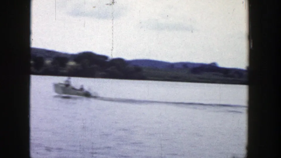 1958 Wisconsin 8mm frame showing a small motorboat crossing calm lake water with a wooded, hilly shoreline in the distance.
