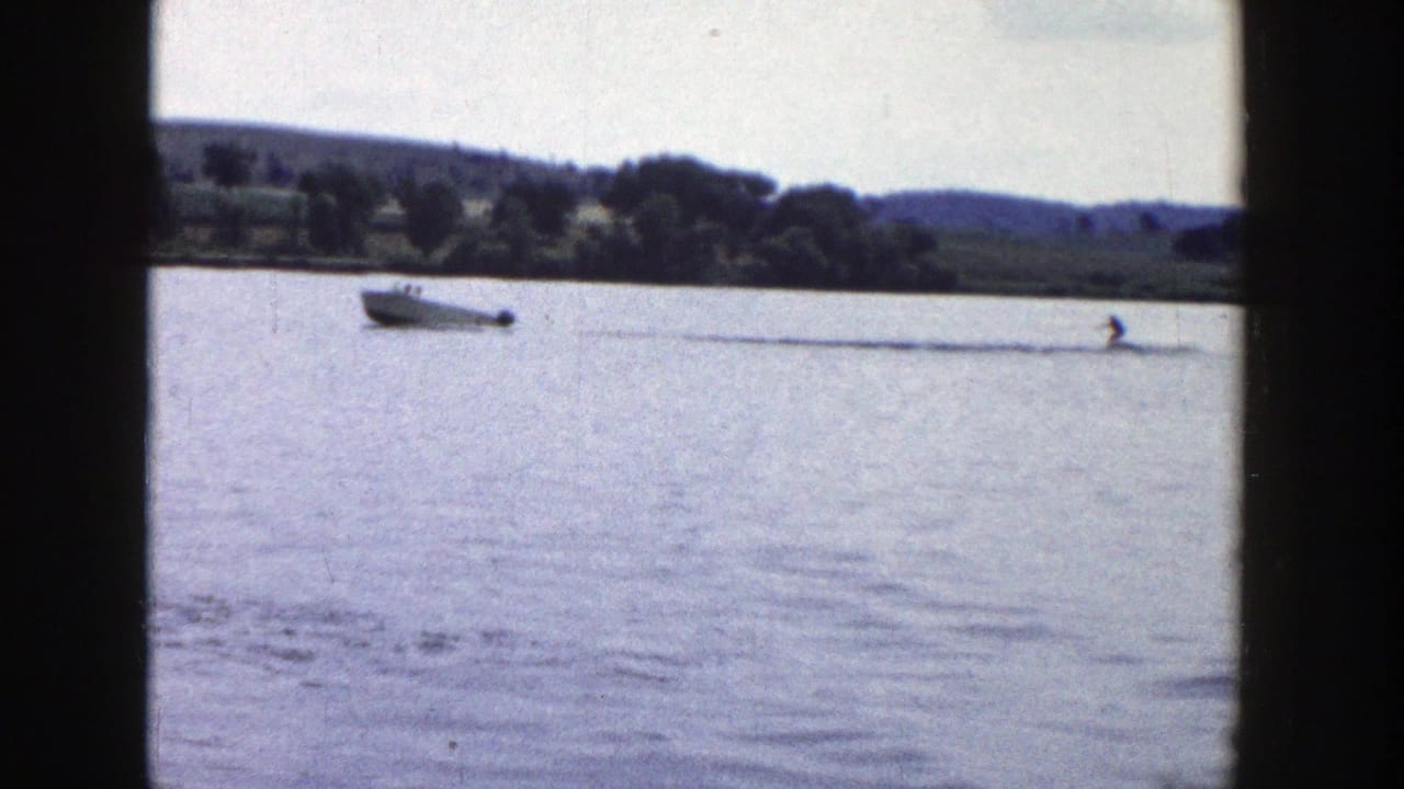 A small motorboat pulls a distant rider across a Wisconsin lake, with calm water, shoreline trees, and hills visible behind.