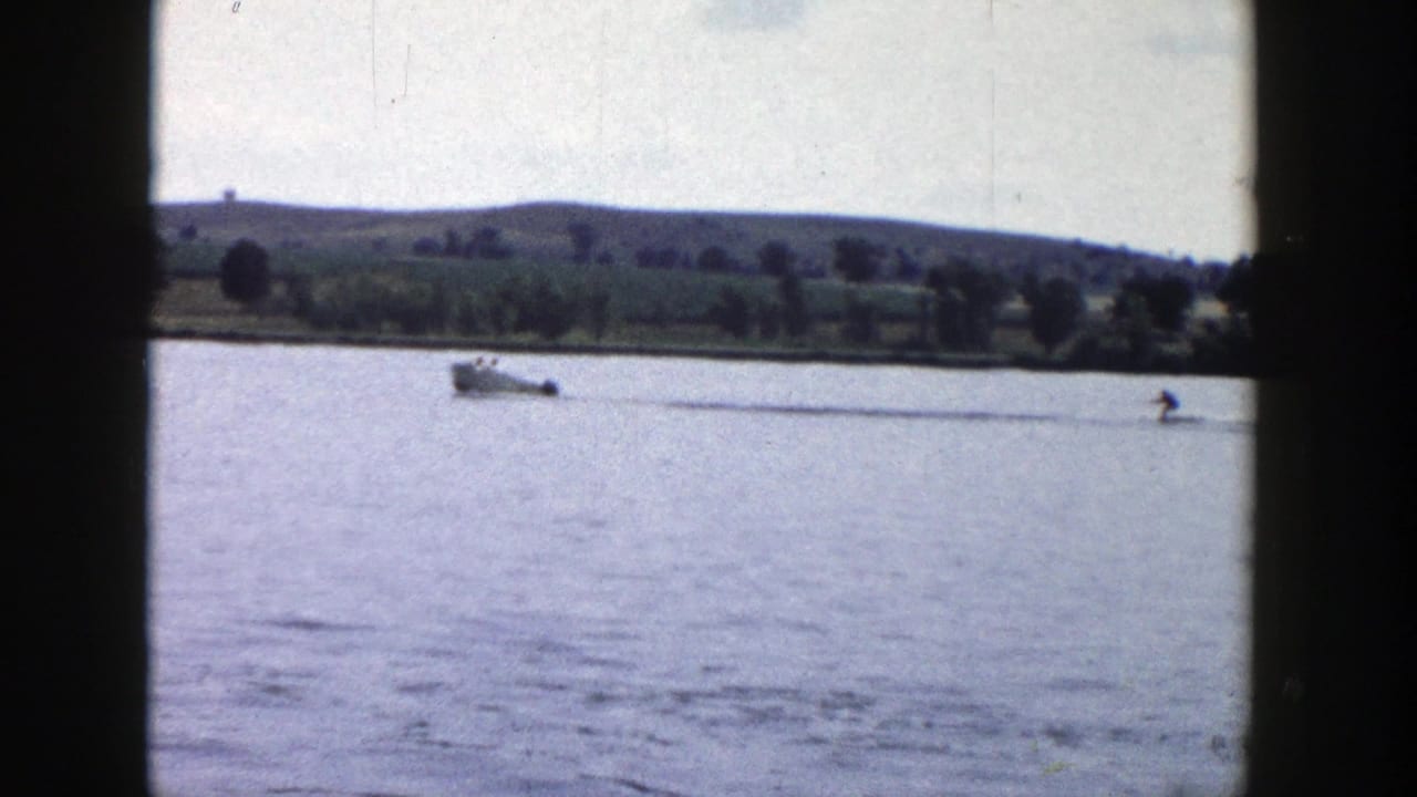 A small motorboat pulls a distant person on a rope across a Wisconsin lake, framed by dark film edges and a hilly shoreline.