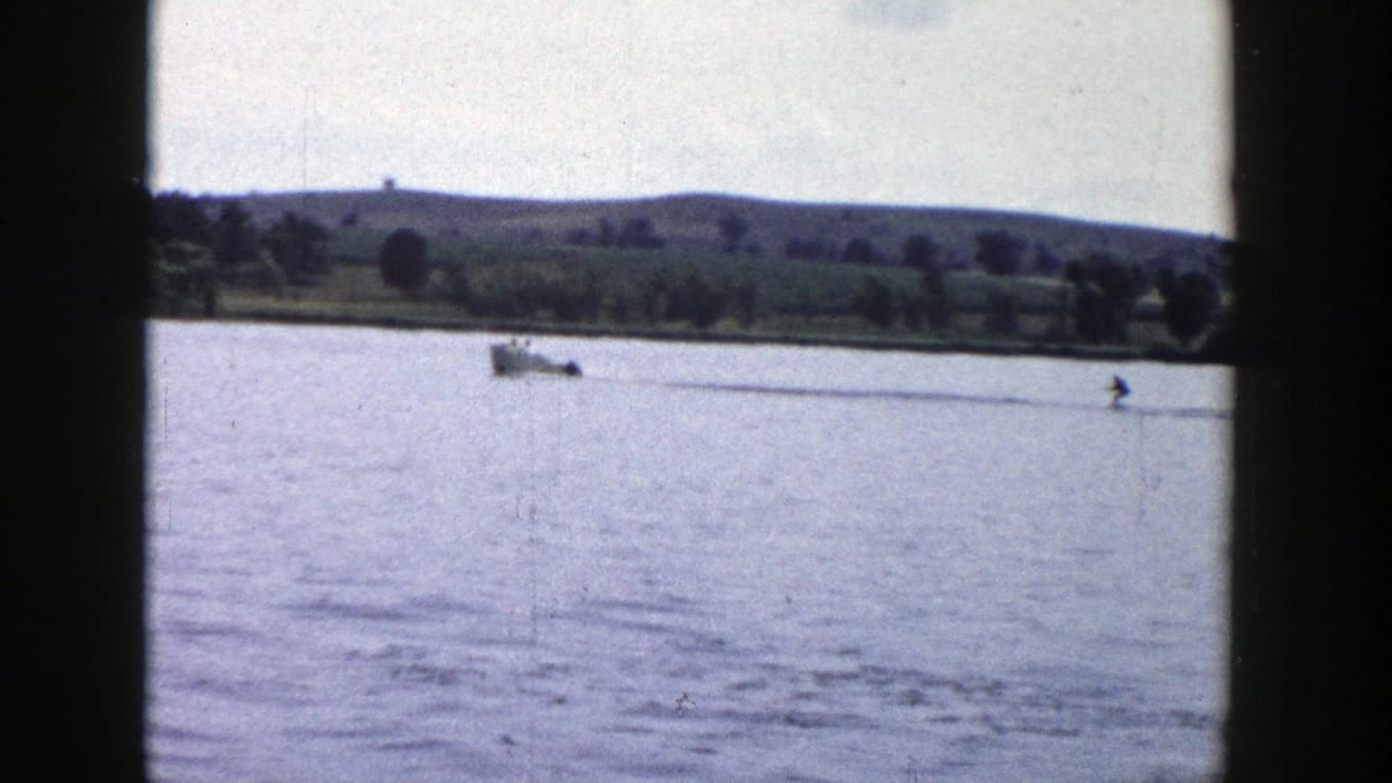 Motorboat towing a distant water skier across a lake with hills and trees beyond.