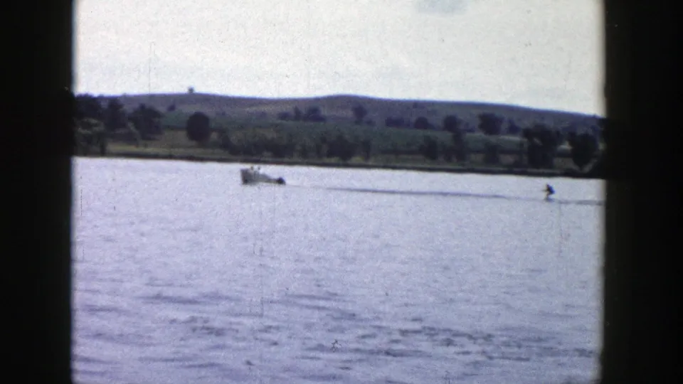 1958 Wisconsin 8mm frame of a motorboat pulling a distant water skier across calm lake water near a rural shoreline.