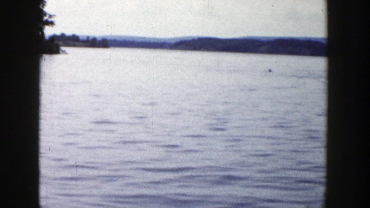 A calm Wisconsin lake fills the frame, with distant hills and a tiny waterborne figure or object near the horizon.
