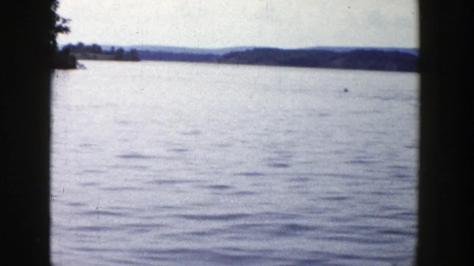 A calm Wisconsin lake fills the frame, with distant hills and a tiny waterborne figure or object near the horizon.