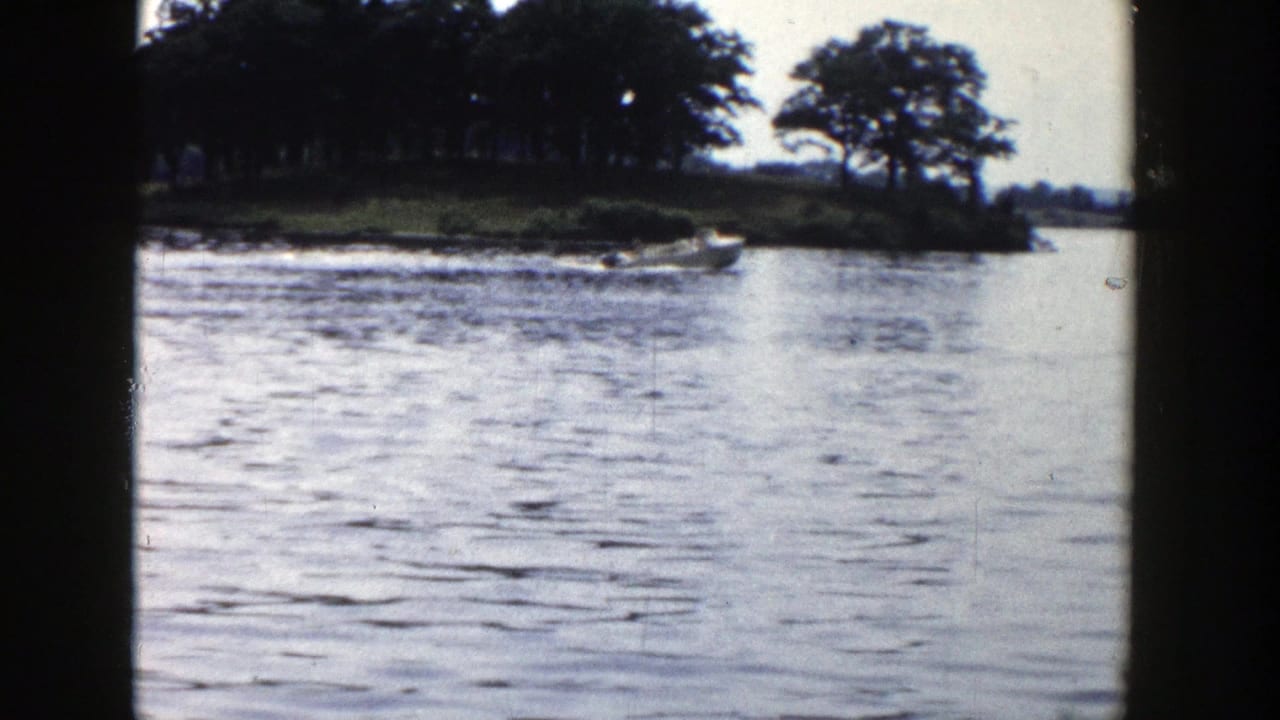 Small boat moving along a Wisconsin lake shoreline in 1958 home-movie footage, with wake and trees behind it.