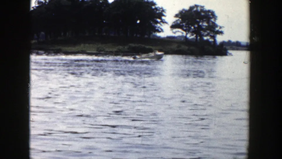 Small boat moving along a Wisconsin lake shoreline in 1958 home-movie footage, with wake and trees behind it.