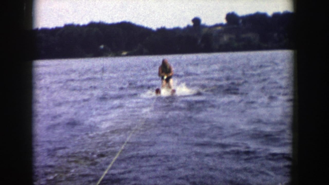1958 Wisconsin home-movie frame of a man water skiing behind a boat on a lake with a tree-lined shore in the distance.