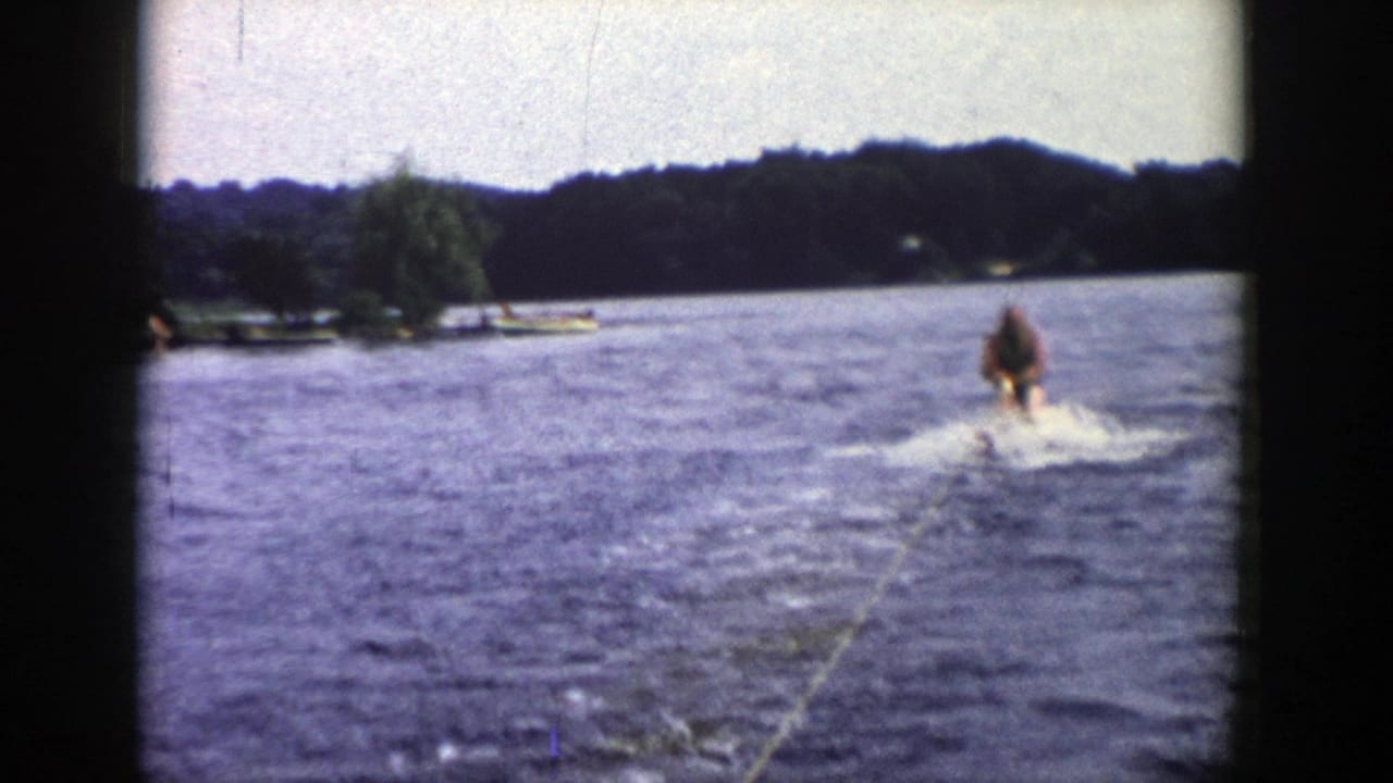 From the moving boat, the 1958 Wisconsin footage shows the skier off to the right with shoreline trees and small lakeside structures in view.