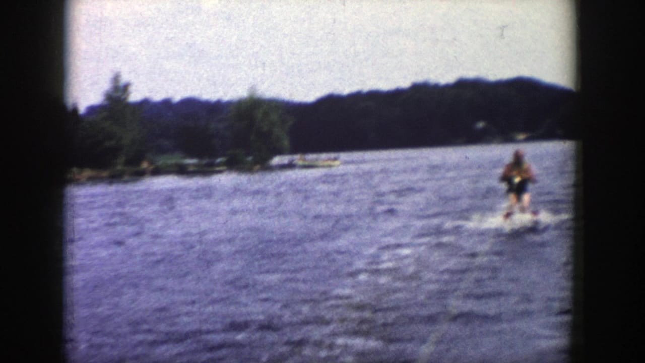 The skier continues upright behind the boat in grainy 8mm footage, with lake wake and a wooded Wisconsin shore behind him.