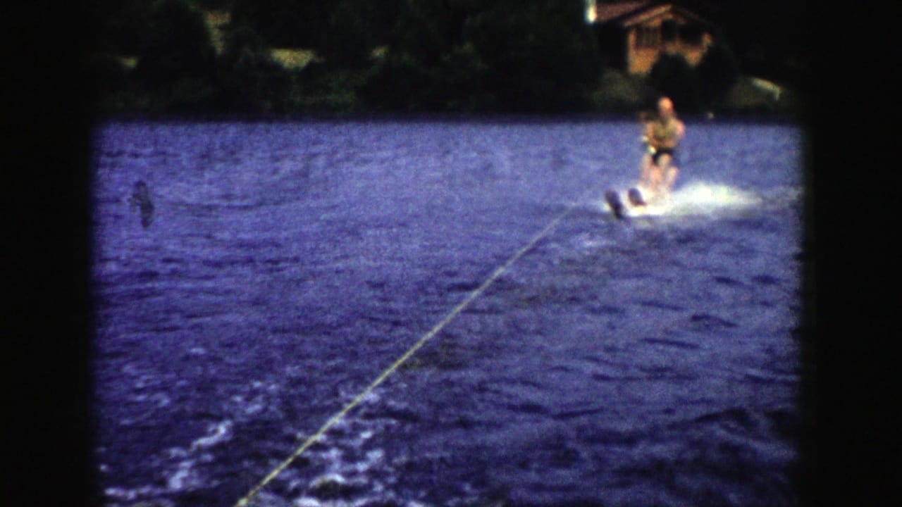 1958 Wisconsin home-movie frame showing a man water skiing across a lake behind an unseen boat, framed by trees on shore.