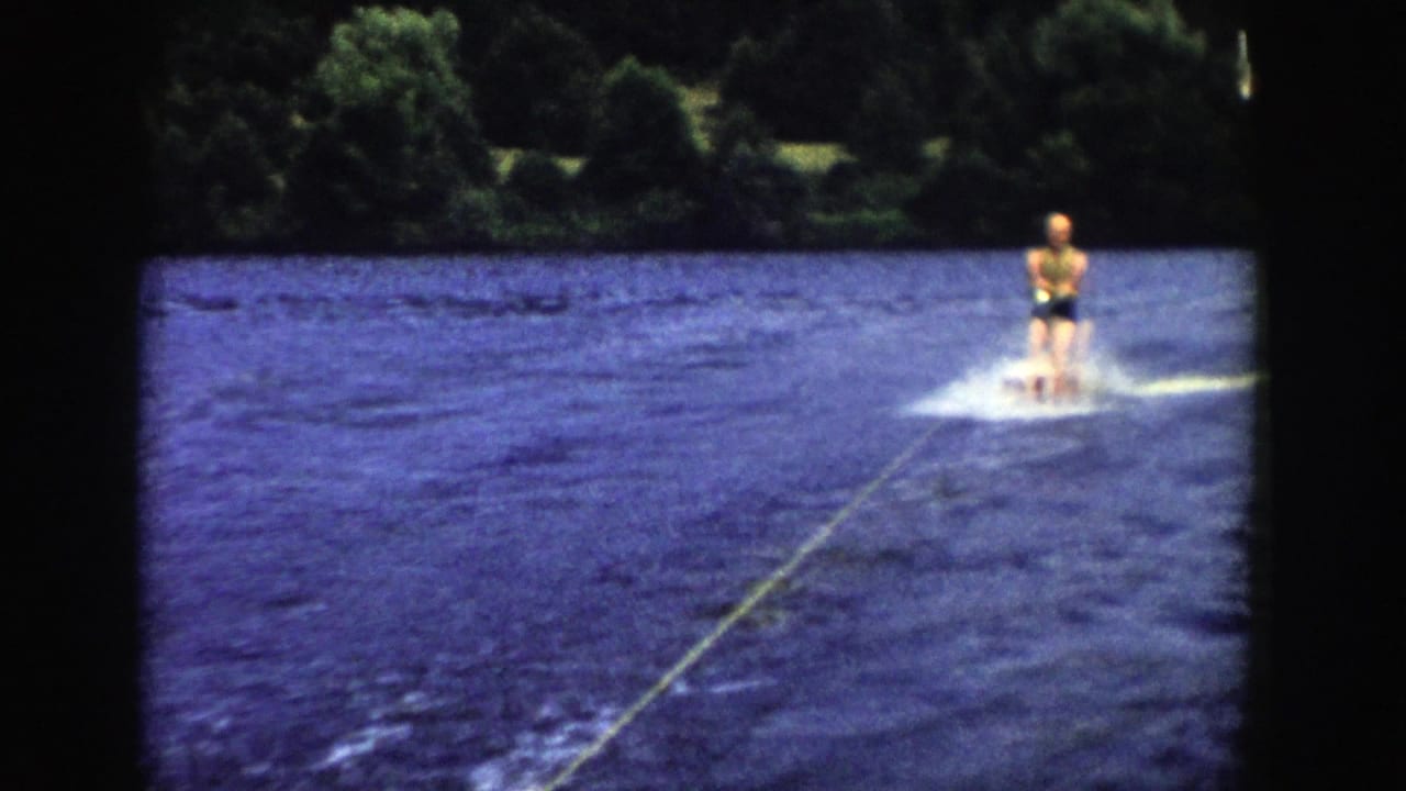 The skier moves farther across the Wisconsin lake, leaving a small wake while dense trees line the far shore.