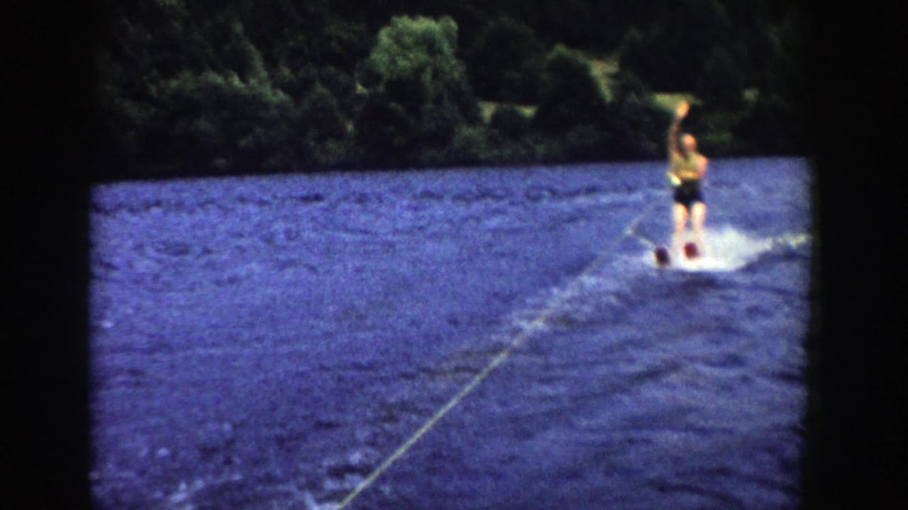 A man in light top and dark trunks rides water skis near the edge of the frame in this 1958 Wisconsin lake scene.