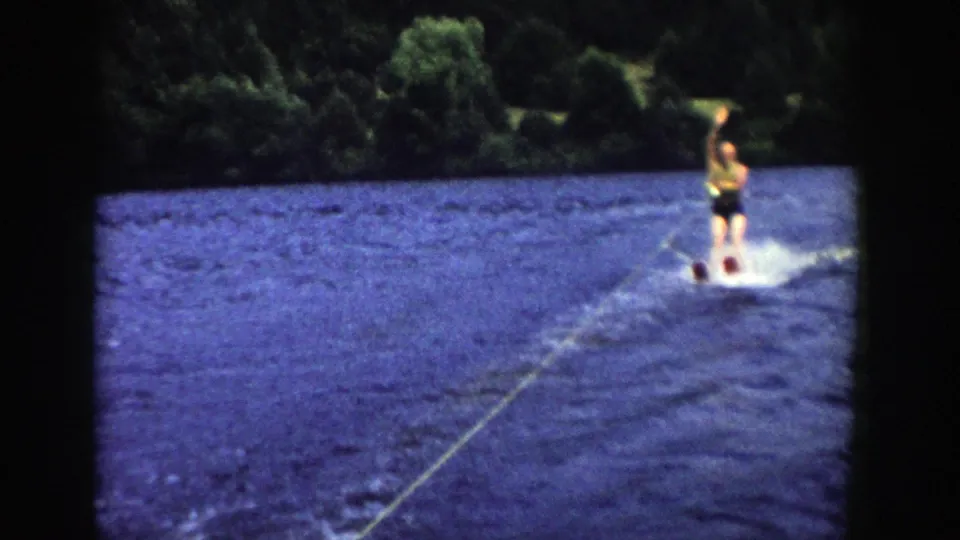 A man in light top and dark trunks rides water skis near the edge of the frame in this 1958 Wisconsin lake scene.