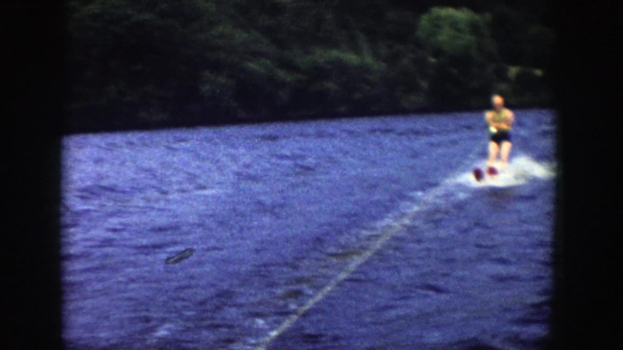 The skier continues across the lake at frame right, with the tow rope and wake visible in soft 8mm color.