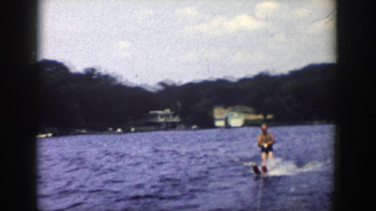 1958 Wisconsin 8mm frame of a young man water skiing on a lake, with trees, lakeside buildings and cloudy sky in the distance.
